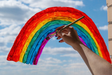 a young girl draws a rainbow on the window glass with a brush and gouache against a blue sky with...