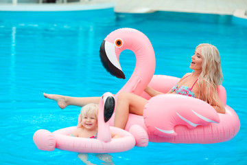 Happy family look summer vacations. Beautiful mother having fun with daughter.  Blonde pretty woman with her little girl wears in fashion same swimwears relax on pink Inflatable pool float flamingo.