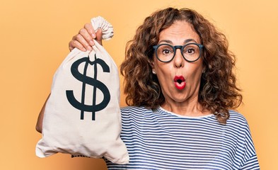 Middle age beautiful woman holding bag with dollar symbol over isolated yellow background scared...