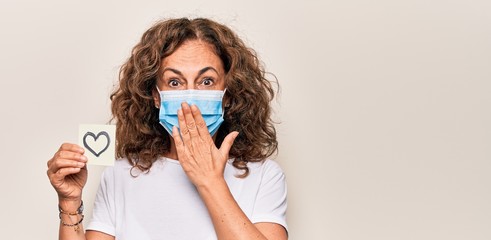 Middle age woman wearing coronavirus protection mask and holding love reminder on paper note covering mouth with hand, shocked and afraid for mistake. Surprised expression