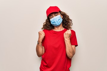 Middle age delivery woman wearing red cap and coronavirus protection mask celebrating surprised and amazed for success with arms raised and eyes closed
