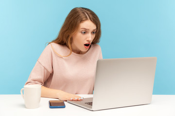 Fototapeta premium Wow, unbelievable! Young woman with shocked expression looking at laptop screen with open mouth, talking on video call, online conference from home office. indoor studio shot isolated, blue background