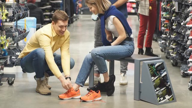 Young beautiful cute caucasian couple in sportswear store, handsome guy sit tying sneakers on his girlfriend.