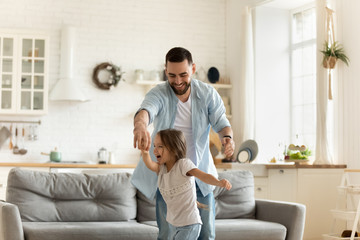 Active young father holding hands of cute little preschool kid daughter, dancing to energetic pop music in modern living room. Happy different generations family having fun together at home.