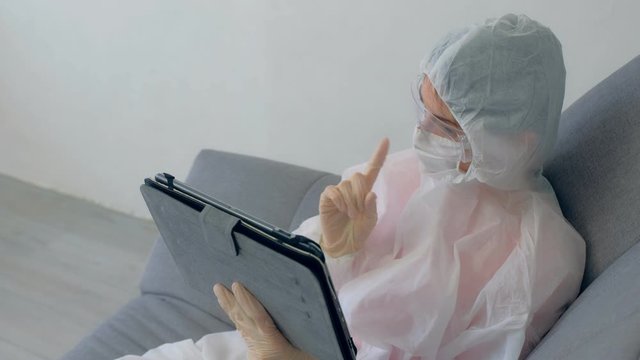 Woman hypochondriac in protective suit, mask, glasses, gloves talking video chat on tablet sitting on couch at home in coronavirus quarantine on pandemic. COVID-19 epidemic, 2019-ncov pandemic.