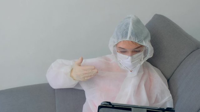 Woman hypochondriac in protective suit, mask, glasses, gloves talking video chat on tablet sitting on couch at home in coronavirus quarantine on pandemic. COVID-19 epidemic, 2019-ncov pandemic.