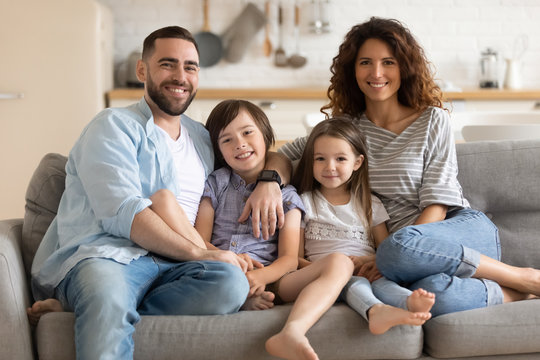 Portrait of happy married couple embracing little kids son daughter, sitting together on comfortable couch in modern studio living room. Smiling cheerful spouses posing for photo with children.