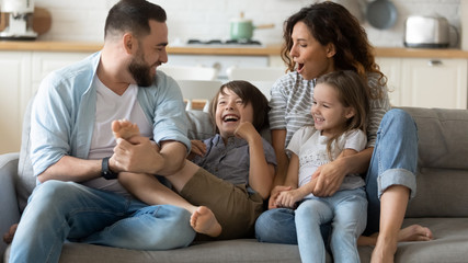 Obraz na płótnie Canvas Happy young father tickling little kid boy son while smiling mother cuddling small daughter on couch. Cheerful full family of four having fun on sofa, enjoying weekend holiday time together at home.