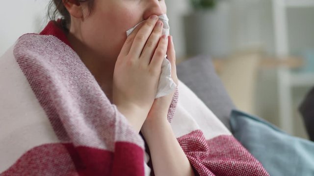 Midsection shot of sick woman with blanket on shoulders covering her mouth with napkin and coughing while sitting on couch at home