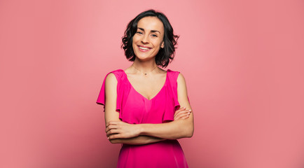 Fototapeta premium True lady. Close-up photo of a charming healthy woman in a fuchsia dress, who is looking in the camera and smiling, while standing with her arms folded.