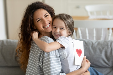 Head shot close up smiling cute little kid girl embracing young woman mother babysitter, congratulating with birthday or special event, preparing handmade greetings post card, looking at camera.