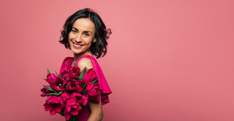 Tender bouquet. Close-up photo of a brunette woman dressed in pink, who is holding a bouquet of tulips and smiling happily.