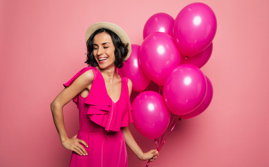 Her perfect birthday. Young charming woman in a fuchsia dress and straw hat is holding pink balloons in her left hand and laughing.