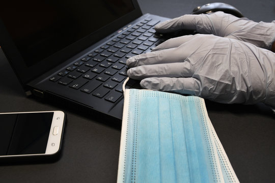 man working on laptop and wearing latex gloves for virus prevention. Man working from home using smartphone and laptop. Surgical face blue masks for coronavirus, covid-19 protection.