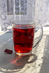 A transparent cup of tea illuminated by the sun's rays from the window is on the table opposite the window, decorated with lace curtains