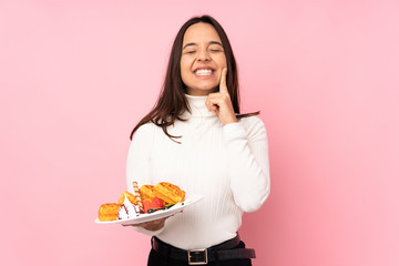 Young brunette woman holding waffles over isolated pink background smiling with a happy and pleasant expression