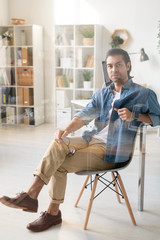 Portrait of young brunette man in casual outfit sitting with eyeglasses in cozy office, behind glass view