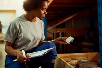 Black female worker taking notes while checking metal tiles in a distribution warehouse.