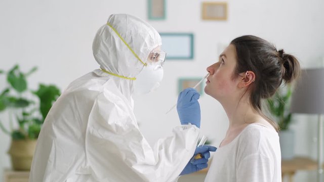 Side view of professional healthcare worker in protective uniform walking to young woman and taking nasal swab from her nose while doing covid test at home