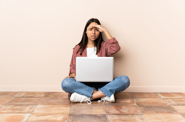 Young mixed race woman with a laptop sitting on the floor looking far away with hand to look something