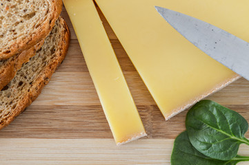 slice of french comte cheese on wood cutting board with fresh green spinach leaves and sliced 7-grain bread closeup