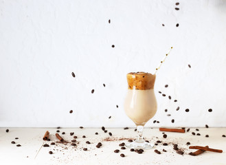Dalgona coffee, a modern korean smooth, creamy and whisked coffee with milk and ice with cinnamon sticks with streaming cohhee beans. Healthy cocktail with soy milk and carob in glass with paper straw