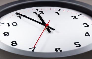 It is five to twelve, the clock is ticking. Modern white grey clock shows the time 5 before 12. Close up to a wall clock, with five minutes to twelve o'clock. Time is running out 