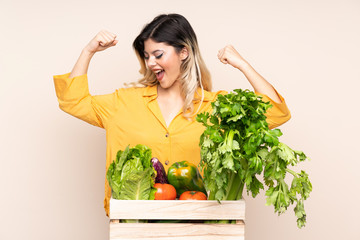 Teenager farmer girl with freshly picked vegetables in a box isolated on beige background celebrating a victory