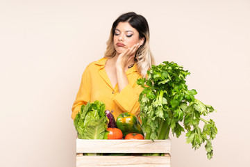 Teenager farmer girl with freshly picked vegetables in a box isolated on beige background unhappy and frustrated