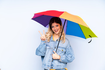 Teenager russian girl holding an umbrella isolated on blue background smiling and showing victory sign