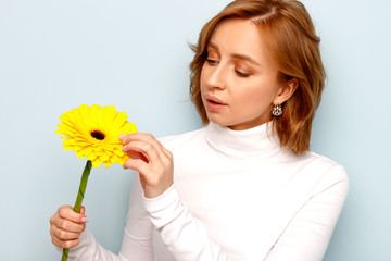 Young woman in white turtleneck holding and admires yellow gerbera flower, fingers touch the petals, isolated on blue background. 
