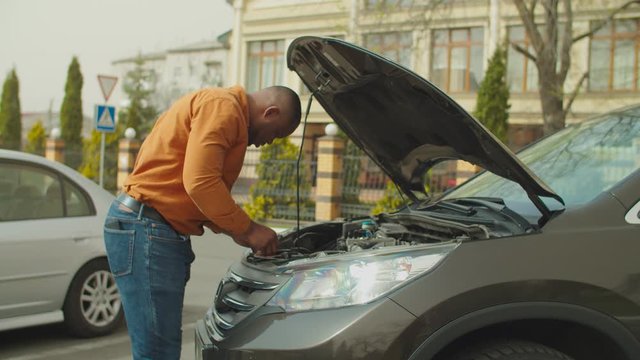 Upset handsome african male driver in casual clothes checking and trying to fix broken down car engine on the street during road trip. Black man looking at broken engine under car hood outdoors.