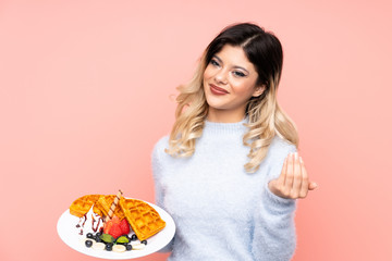 Teenager girl holding waffles on isolated pink background inviting to come with hand. Happy that you came