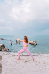 Pregnant woman practices yoga, standing in Virabhadra position on the beach. Woman with tattoo in pink and purple tracksuit. Calm girl practicing at dawn, meditation on the sandon the sand.