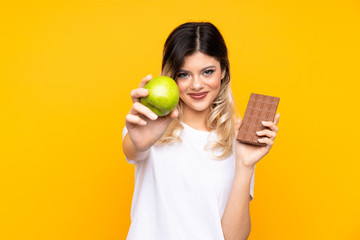 Teenager girl isolated on  yellow background taking a chocolate tablet in one hand and an apple in...