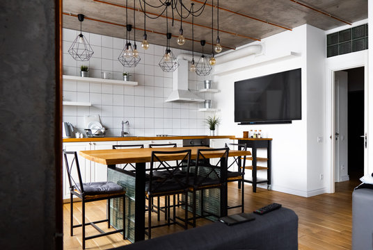 An eat-in kitchen interior design in modern scandinavian style with big wooden table and chairs against light wood floor, bright white walls and furnitures with TV, appliances and hanging light bulbs