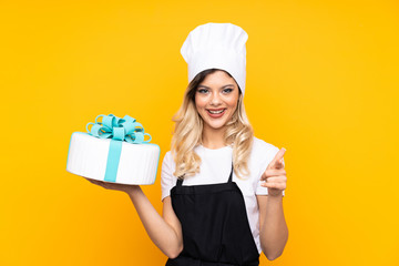 Teenager girl pastry holding a big cake isolated on yellow background points finger at you