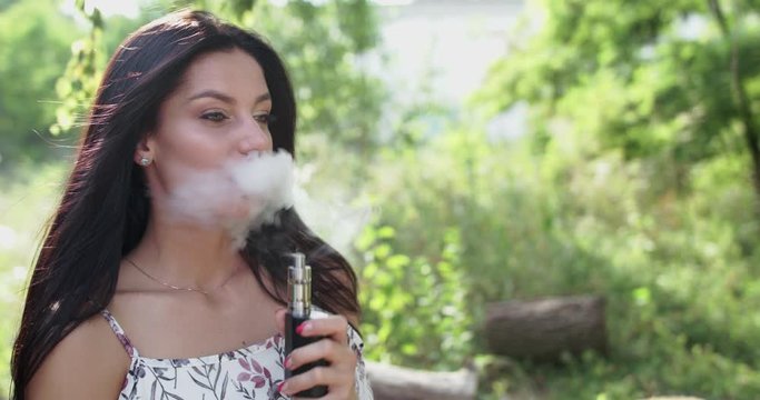 Portrait of pretty brunette smoking e-cigarette and exhales at camera on nature