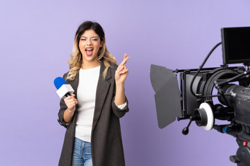 Reporter teenager girl holding a microphone and reporting news isolated on purple background with fingers crossing