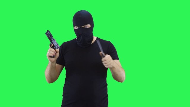 A man in a black mask holds a knife and a gun in his hand.Balaclava.Green screen background.