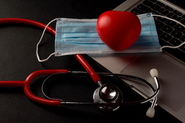 Stethoscope, face mask and red heart with laptop on dark background. Health concept. Selective...