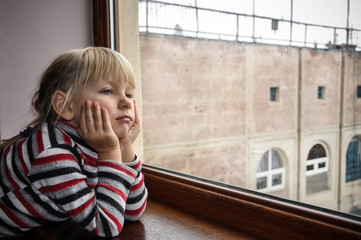 White-haired girl looks out the window. Quarantined child. The child is sad at the window.