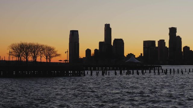 View of Jersey city skyline silhouette at sunset from the Hudson River boardwalk with a small pier with a tree in an orange backlight on the left of the image