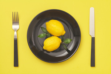 Black plate, knife and fork with lemon on yellow background. Top view.