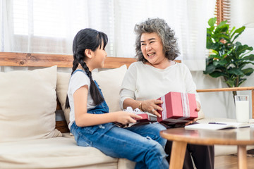 Asian young little girl give gift to grandmother at home. They sit on sofa together. Old smile happy woman open box with granddaughter. Two generation have happiness moment. Family lifestyle concept.