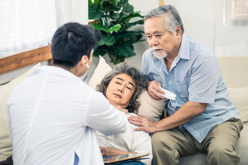 Asian senior old woman with bad condition feeling sick lying on sofa having medical check up by doctor visiting at home. Elder man husband taking care of wife closely and worry about unwell illness.