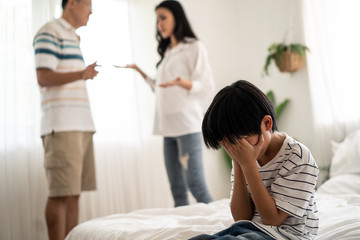 Obraz premium Asian boy kid sitting and crying on bed while parents having fighting or quarrel conflict at home. Child covering face and eyes with hands do not want to see the violence. Domestic problem in family.