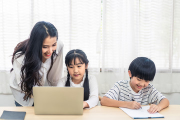 Asian parent teaching homework to children after learning online education class from school teacher at home. Mother explaining knowledge in computer laptop to daughter and helping son lecture notes.