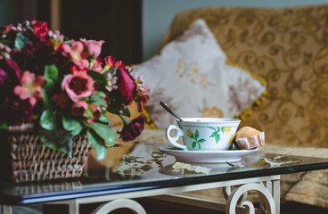 Cup of tea with cupcake next to flowers and sofa background
