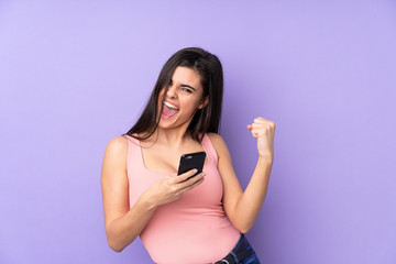 Young woman over isolated purple background with phone in victory position